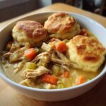A white bowl filled with creamy chicken and dumpling stew topped with three golden brown biscuits.
