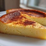 A close-up of a thick slice of creamy yellow egg custard with a deeply browned, spiced top, served on a white plate.