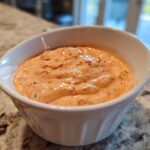 A close-up of creamy, orange-hued burger sauce speckled with herbs and spices, served in a small white bowl.