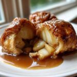 A close-up view of a warm apple dumpling cut in half, revealing soft apple filling, covered in glossy caramel sauce.