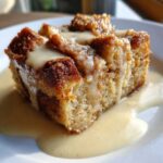 A close-up of a moist slice of bread pudding topped with creamy vanilla sauce on a white plate.