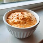 A close-up of creamy, orange-colored burger sauce speckled with red pieces in a small white bowl.