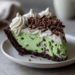 A close-up slice of amazing grasshopper pie featuring a mint-green filling with chocolate chips, a dark crust, and whipped cream topping.