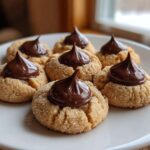 Six freshly baked peanut butter blossoms, rolled in sugar and topped with a chocolate kiss, arranged on a white plate.