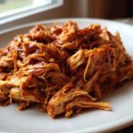 A close-up of saucy, shredded pulled chicken piled high on a white plate.