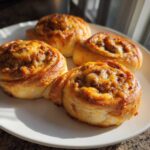 Four freshly baked, golden brown sausage pinwheels topped with melted cheese resting on a white plate.