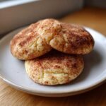 Three freshly baked, soft snickerdoodle cookies generously coated in cinnamon sugar, stacked on a light gray plate.