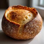 Close-up of a freshly baked artisan bread bowl with a thick, golden-brown, flour-dusted crust, ready to be filled.