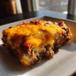 A close-up, appetizing slice of bacon cheeseburger casserole topped with melted cheddar cheese.