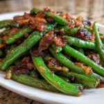 A close-up of bright green beans tossed with crispy, caramelized bacon bits served on a white platter.
