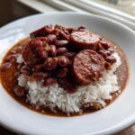 A white bowl filled with white rice topped with savory red beans and thick slices of smoked sausage, a perfect serving of beans and rice.