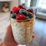 A hand holding a mason jar filled with creamy overnight oats, topped generously with fresh raspberries, blueberries, and seeds.