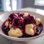 Close-up of fluffy dumplings generously covered in a deep purple blackberry sauce, served in a white bowl.