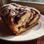 A close-up of a freshly baked chocolate babka loaf, showing rich chocolate swirls and a white glaze drizzle, sitting on a white plate.