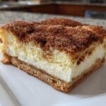 A single, thick slice of churro cheesecake bar featuring a creamy white filling and a heavy cinnamon-sugar topping.