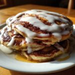 A stack of fluffy cinnamon roll pancakes topped with thick white glaze and syrup on a white plate.