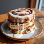 A tall stack of three cinnamon roll pancakes generously drizzled with white cream cheese icing.