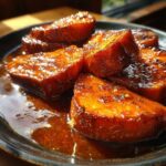 Close-up of thick-cut, glazed candied yams soaking in rich brown syrup on a dark plate.