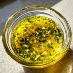 Close-up of bright yellow cowboy butter dipping sauce, rich with minced garlic and green herbs, in a small glass bowl.