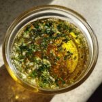 Overhead view of vibrant cowboy butter dipping sauce, rich yellow oil mixed with minced garlic and fresh green herbs in a small glass jar.