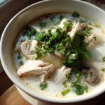 A close-up bowl of creamy chicken coconut soup, topped generously with shredded chicken, mushrooms, and fresh cilantro.