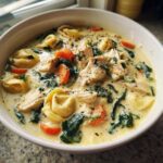 Close-up of a hearty bowl of creamy chicken tortellini soup, featuring chunks of chicken, spinach, and sliced carrots.