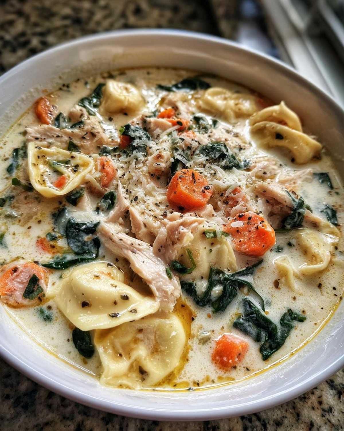 A close-up view of a white bowl filled with creamy chicken tortellini soup, featuring pasta, shredded chicken, carrots, and spinach.