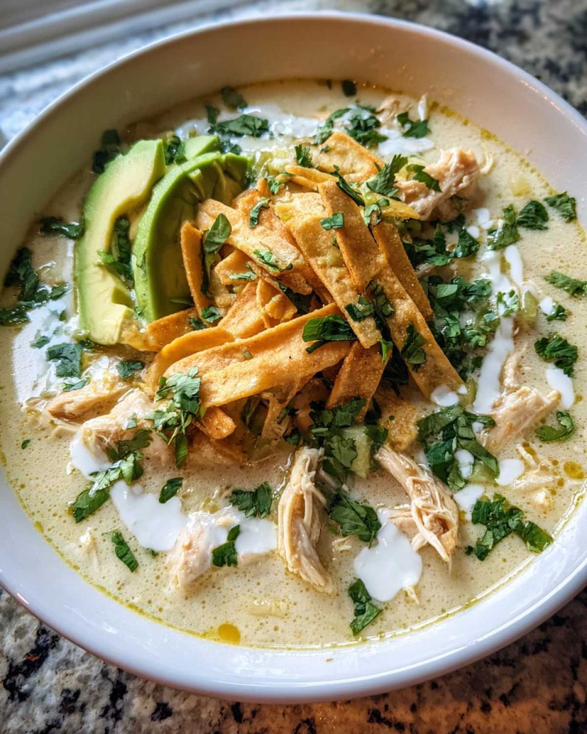 A close-up of a bowl of creamy chicken tortilla soup topped with avocado, tortilla strips, and cilantro.