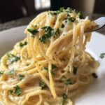 A fork lifts spaghetti coated in rich, creamy lemon pasta sauce, garnished with parsley and pepper.