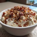 Close-up of a white bowl filled with creamy Neiman Marcus dip, generously topped with crumbled bacon and chopped pecans.