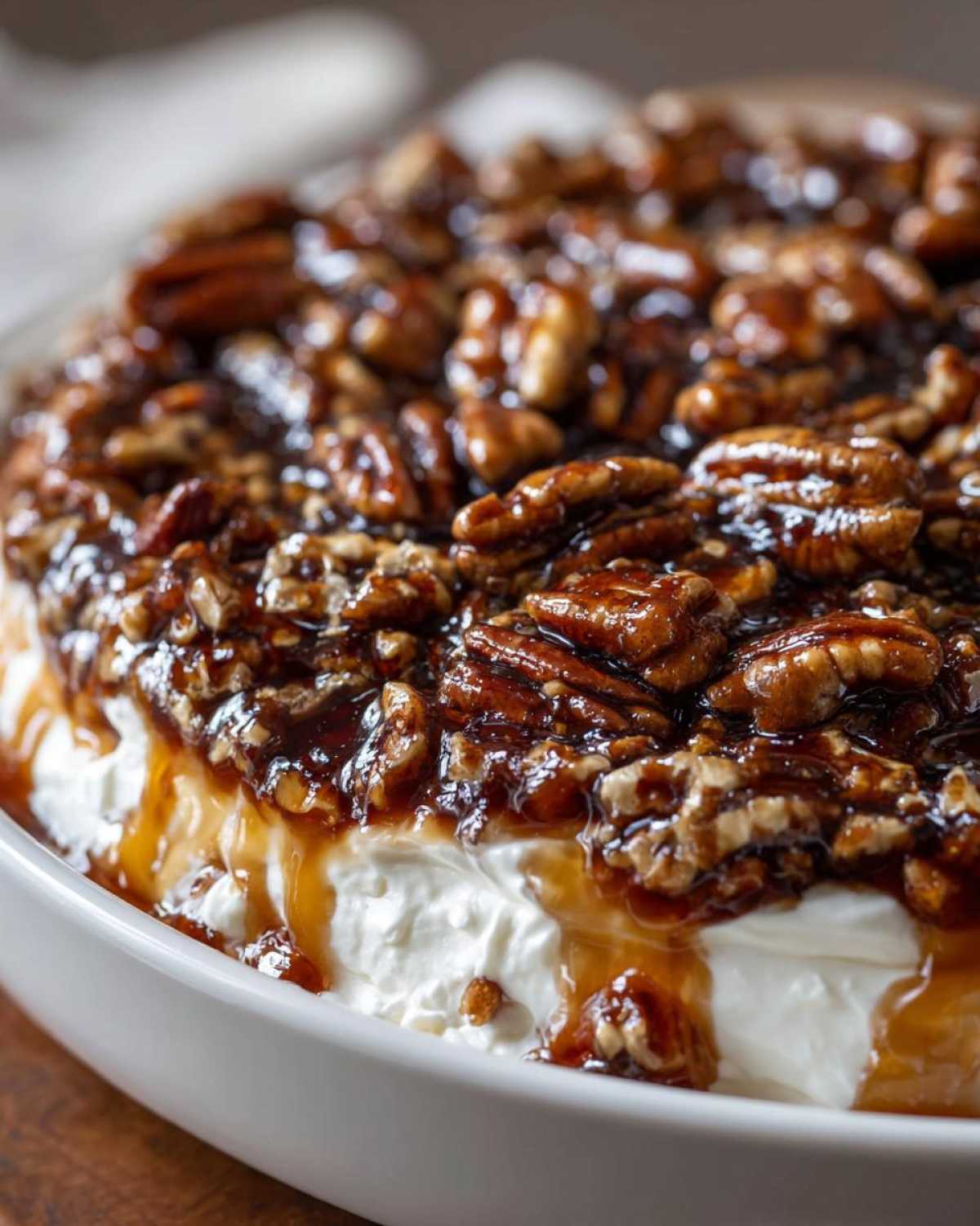 A close-up shot of a creamy pecan pie dip topped generously with glazed pecans and caramel sauce dripping down the sides.