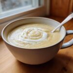 A creamy bowl of roasted cauliflower soup, garnished with a swirl of cream and pepper, sitting on a wooden surface.