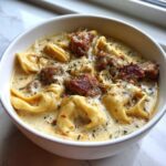 Close-up of a white bowl filled with creamy sausage tortellini soup, topped with herbs and cheese.