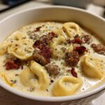 Close-up of a white bowl filled with creamy sausage tortellini soup, topped with crumbled sausage and herbs.