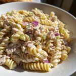 A close-up of creamy tuna pasta salad made with fusilli pasta, red onion, and celery in a white bowl.