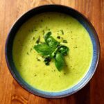Overhead view of a bowl of vibrant green, creamy vegan zucchini soup topped with fresh basil leaves and cracked black pepper.