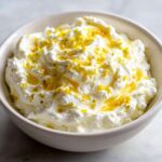Close-up of creamy whipped feta dip in a white bowl, drizzled with oil and topped with bright yellow lemon zest.
