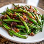 Close-up of bright green beans tossed with crispy, caramelized bacon pieces on a white plate.