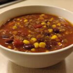 A close-up of a white bowl filled with steaming crockpot cowboy soup, featuring ground beef, kidney beans, and corn.