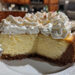 A close-up of a thick slice of eggnog cheesecake topped with whipped cream and sprinkled with cinnamon.