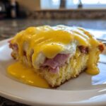 A close-up of a square slice of eggs benedict casserole topped with ham, egg, and dripping hollandaise sauce.
