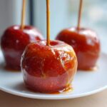 Close-up of a shiny, red caramel apple with hot caramel sauce being drizzled on top, sitting on a white plate.