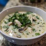 A close-up of a white bowl filled with creamy, savory coconut soup featuring chunks of chicken, mushrooms, and topped with fresh cilantro.