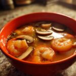 A vibrant orange bowl filled with steaming tom yum soup featuring large shrimp and sliced mushrooms.