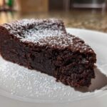 A decadent slice of flourless chocolate torte, dark brown and dense, dusted with white powdered sugar on a white plate.