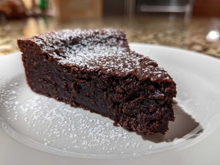 A decadent slice of flourless chocolate torte, dark brown and dense, dusted with white powdered sugar on a white plate.