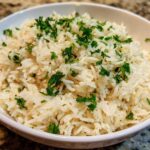 A close-up of fluffy white rice pilaf served in a white bowl and garnished generously with chopped green parsley.