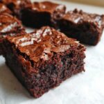 Close-up of a square, fudgy brownie showing its dense interior and signature crackly top, one of several brownies visible.