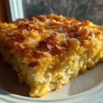 A close-up, sunlit slice of cheesy funeral potatoes recipe with a golden, crunchy cornflake topping.