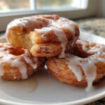 Three golden brown apple fritter donuts stacked on a white plate, heavily drizzled with white glaze.